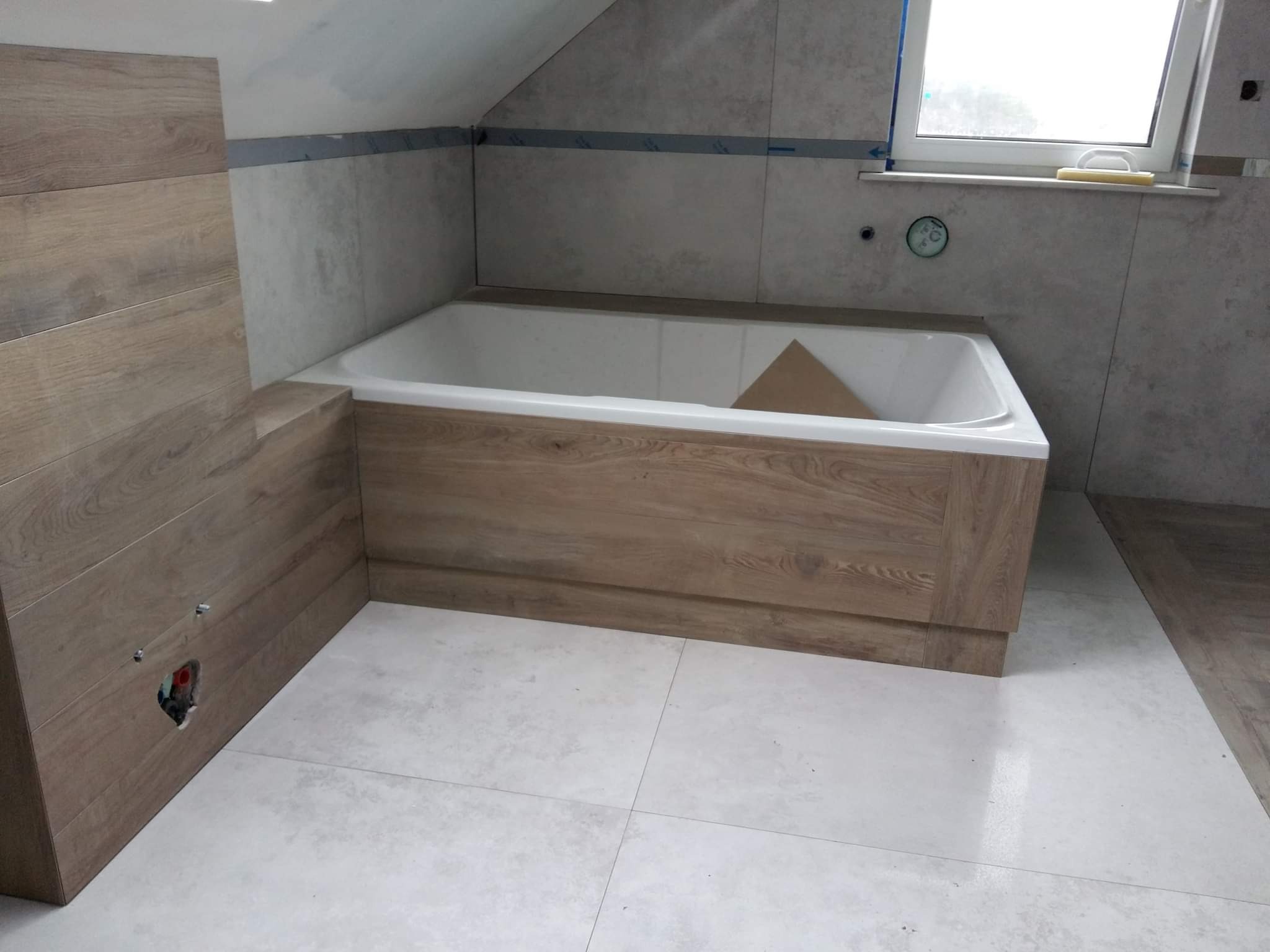 Wykończona łazienka na poddaszu: obudowana wanna z drewnopodobnymi panelami, jasne płytki na podłodze i ścianach, okno dachowe. Widoczny otwór na instalację.