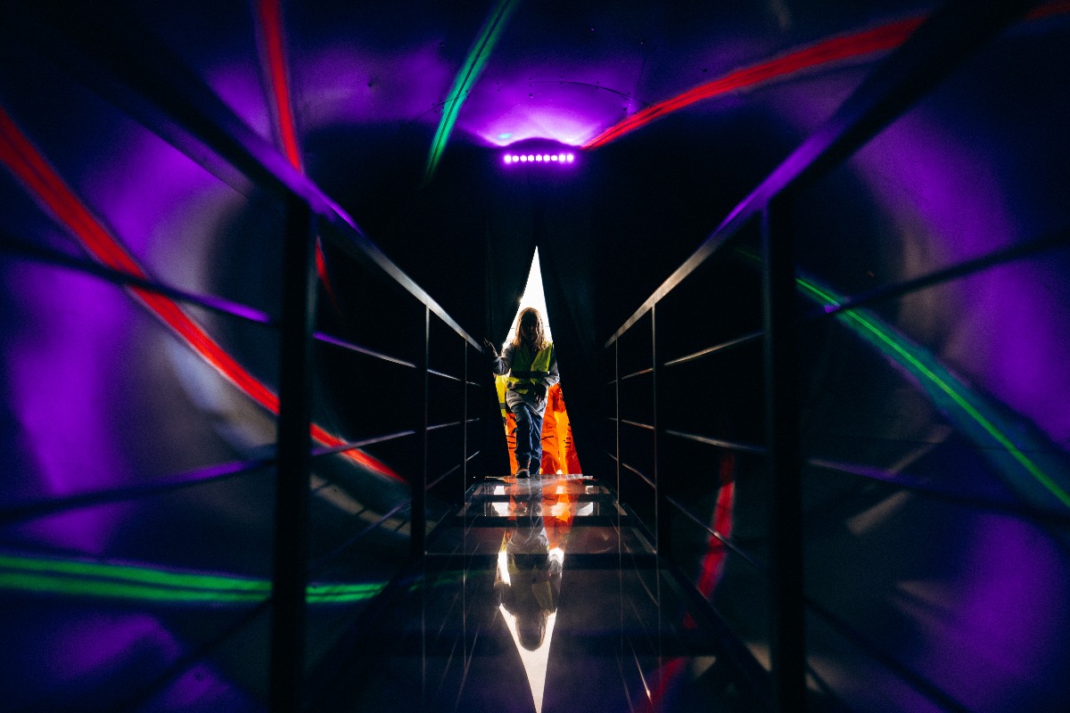 Osoba w kamizelce odblaskowej w tunelu z neonowymi światłami, odbijającymi się w podłodze. U góry rząd fioletowych świateł. Kąt ujęcia z dołu, symetryczny.