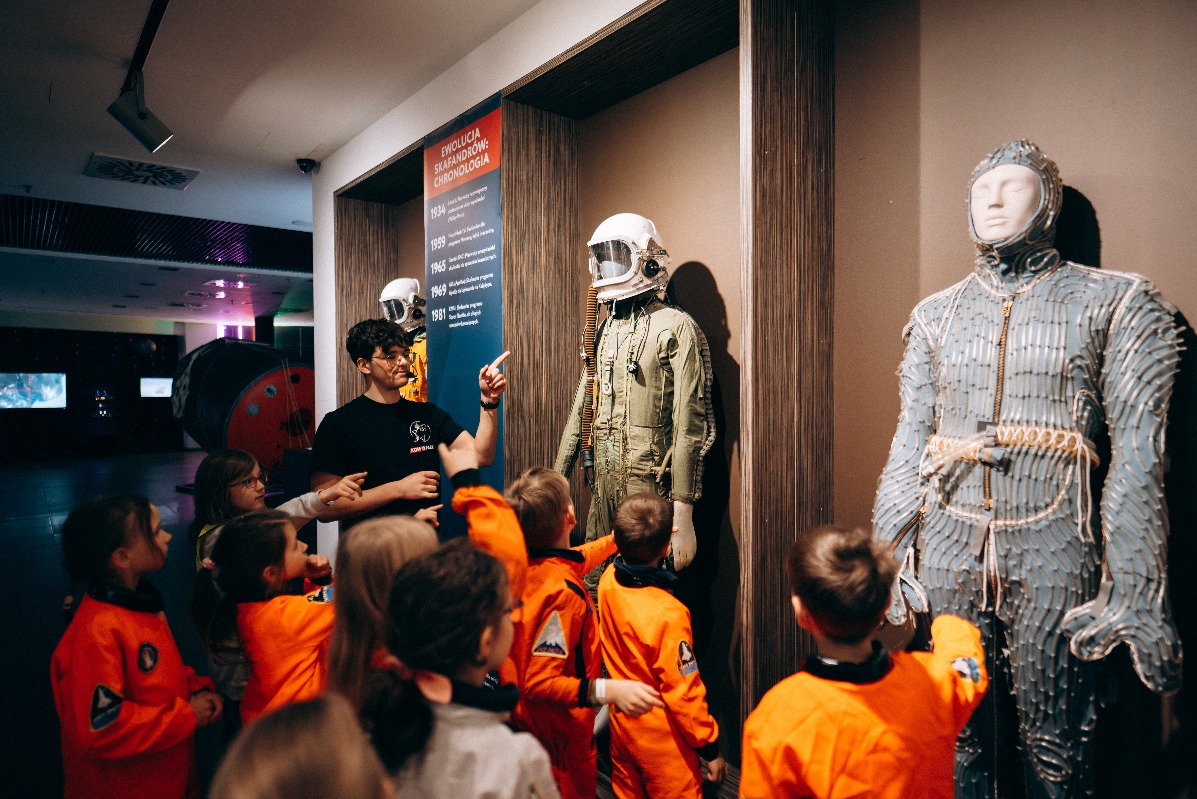 Wycieczka dzieci w pomarańczowych kombinezonach kosmicznych z przewodnikiem w muzeum kosmonautyki, prezentacja skafandrów kosmicznych i ekspozycji o ewolucji skafandrów.