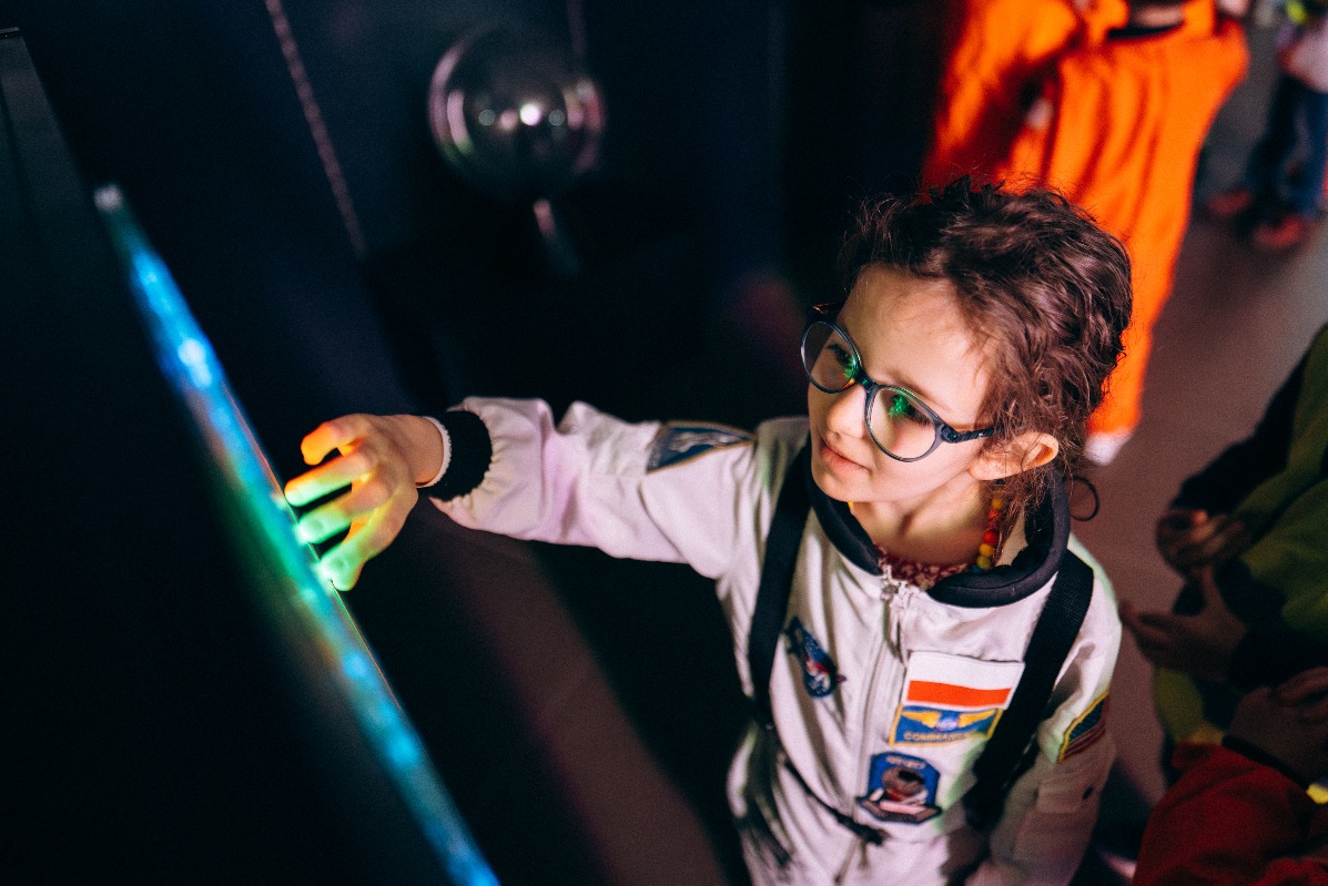 Dziewczynka w kostiumie astronauty dotyka interaktywnego panelu z kolorowymi światłami podczas tematycznego wydarzenia dla dzieci.
