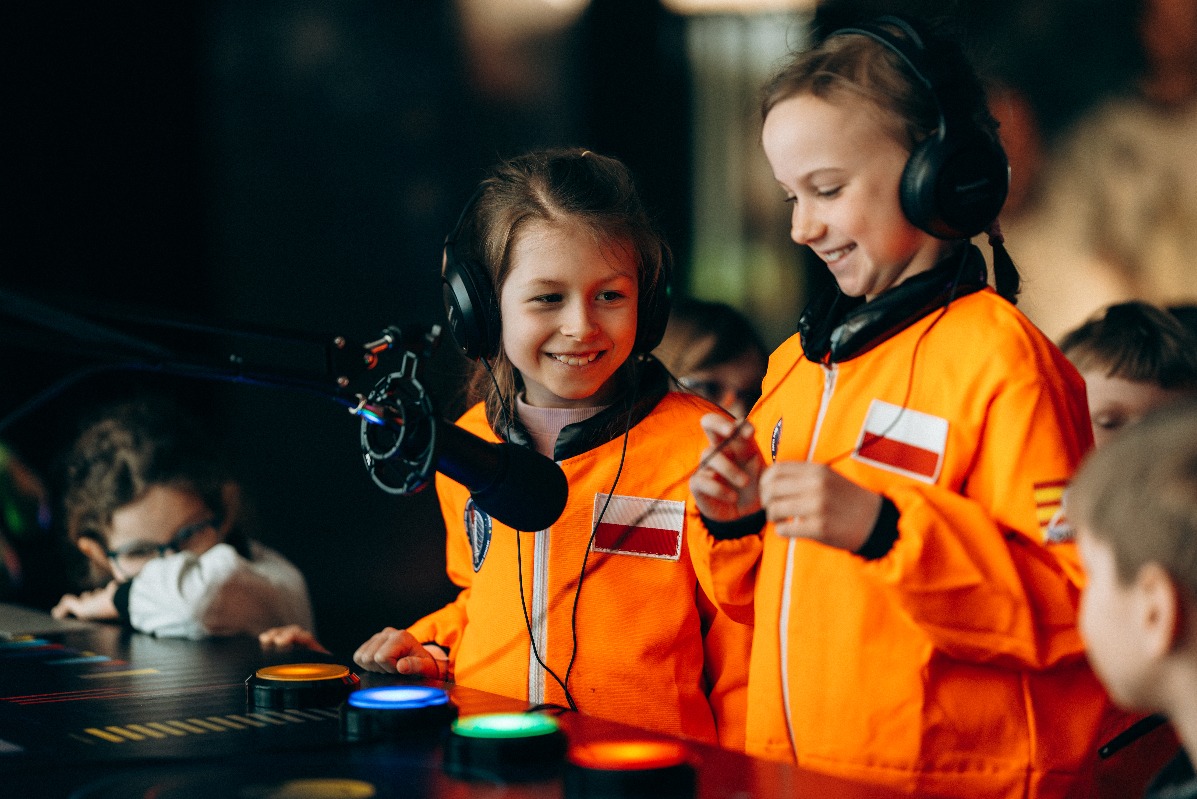 Dzieci w pomarańczowych kombinezonach z flagą Polski, w słuchawkach, bawią się przy interaktywnym panelu z podświetlanymi przyciskami podczas eventu.