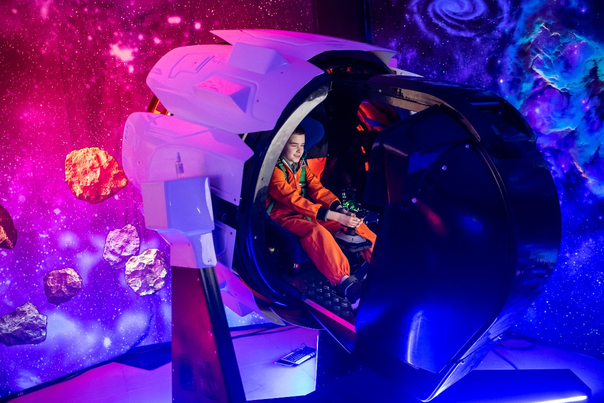 Chłopiec w kombinezonie w symulatorze lotu kosmicznego z tłem galaktyki, oświetlonym neonowym światłem, tworzącym futurystyczną atmosferę.