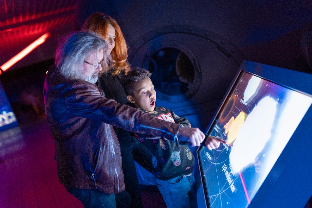 Rodzina zafascynowana interaktywną wystawą w muzeum nauki, chłopiec z otwartymi ustami wskazuje na ekran dotykowy z mapą kosmosu, oświetlenie w odcieniach czerwieni i niebieskiego.