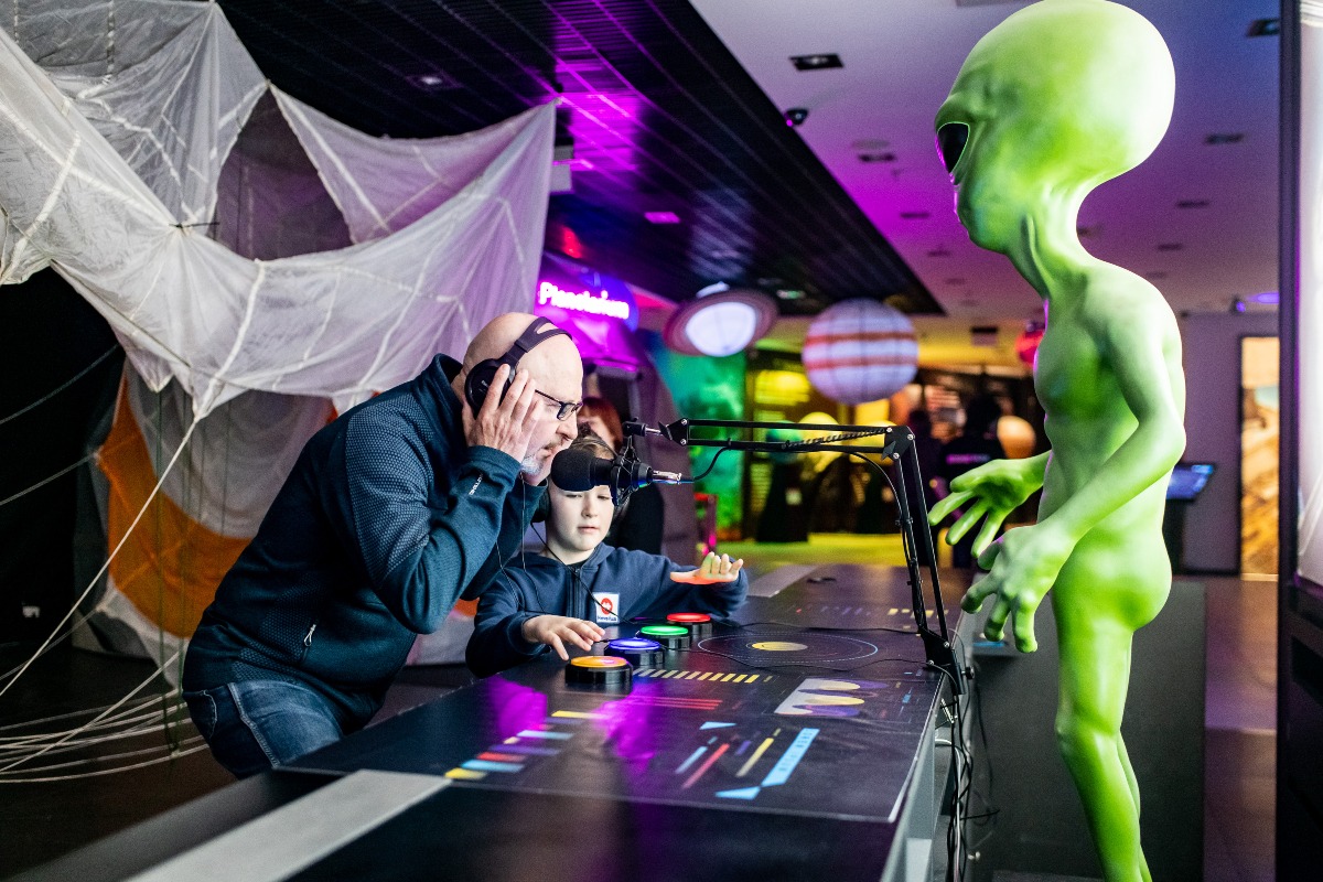Interaktywna wystawa: mężczyzna w słuchawkach i dziecko w VR testują panel sterowania obok figury zielonego kosmity w futurystycznej przestrzeni.