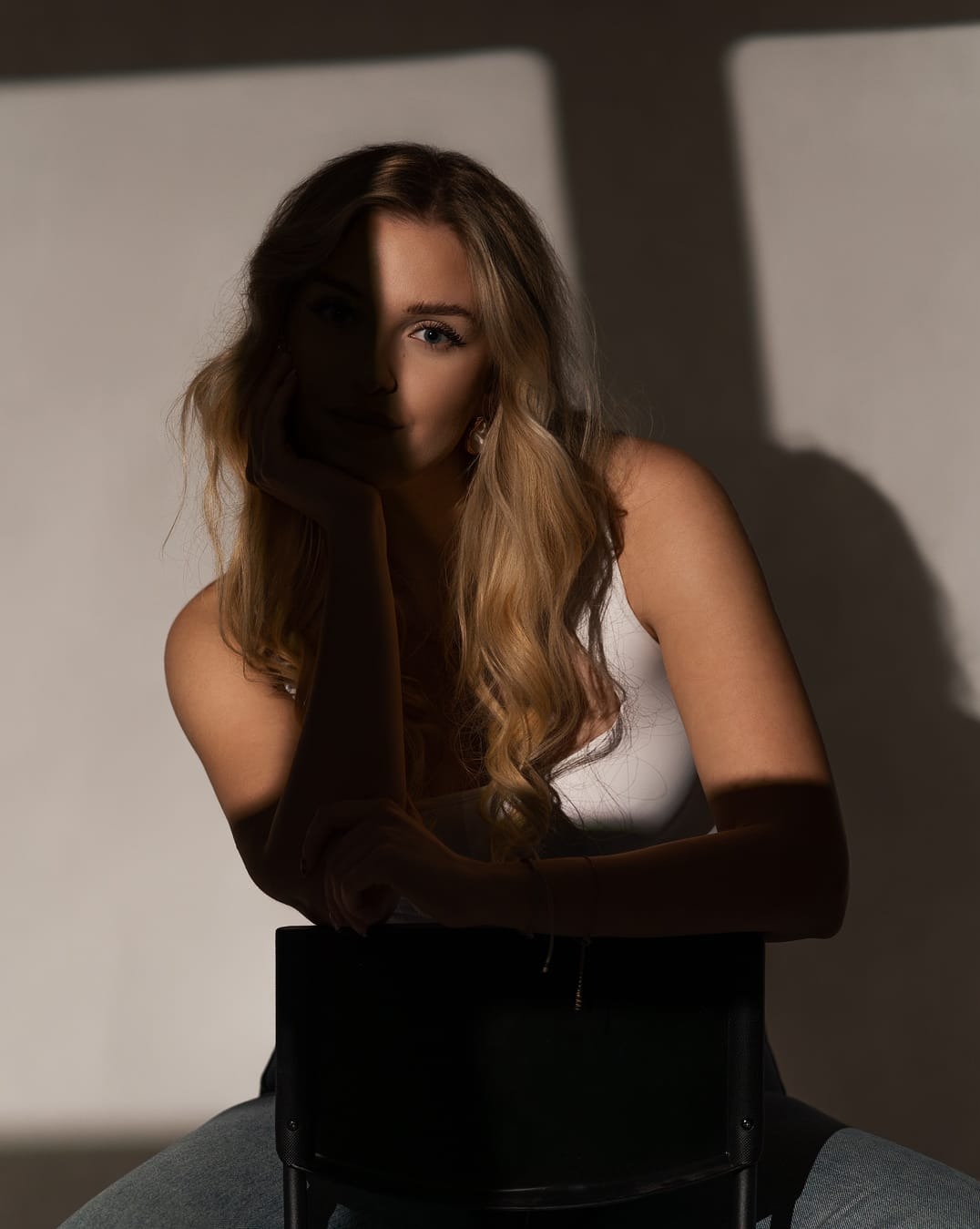 Portret kobiety z blond włosami, oświetlonej kontrastowym światłem, siedzącej na krześle. Cień dzieli twarz na pół, podkreślając niebieskie oczy i subtelny makijaż.