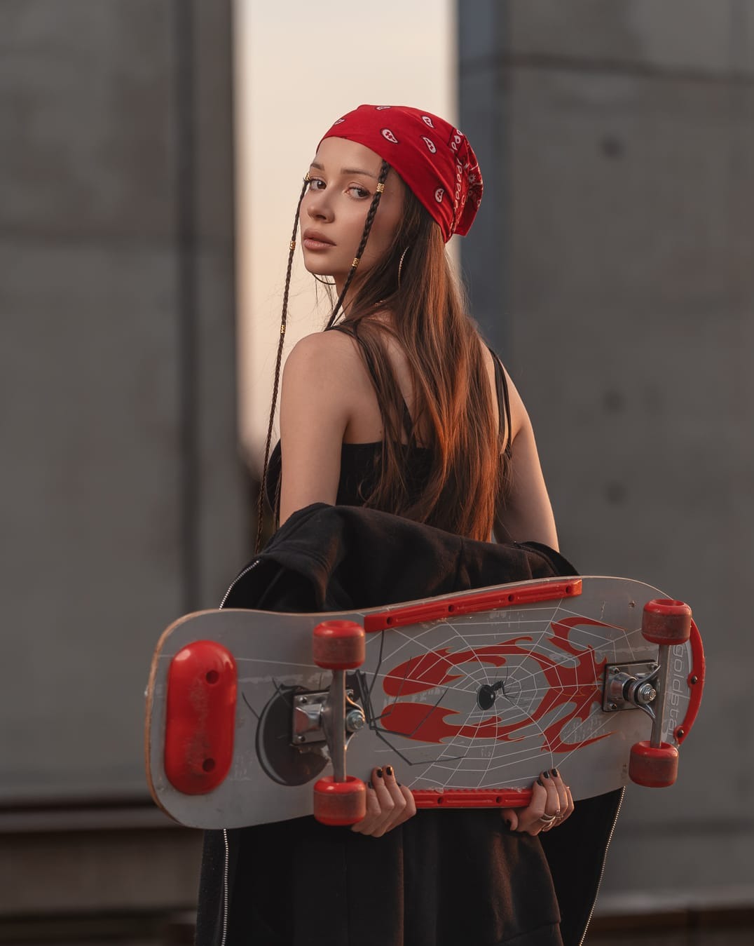 Młoda kobieta z długimi włosami i czerwoną bandaną, odwrócona przez ramię, trzyma deskorolkę na tle betonowej ściany w ciepłym, naturalnym świetle.