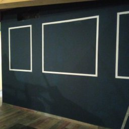 Hubert Słaboń - Ciemna ściana z białymi, geometrycznymi obramowaniami w kształcie prostokątów, drewniany sufit i podłoga. Widoczny fragment drabiny.