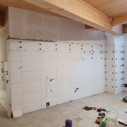 Hubert Słaboń - Białe płytki ścienne z krzyżykami dystansowymi w trakcie montażu, widoczne instalacje i drewniany sufit. Prace wykończeniowe w pomieszczeniu z podłogą w jasnych płytkach.