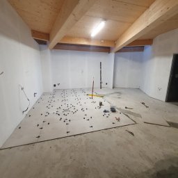 Hubert Słaboń - Układanie płytek podłogowych w surowym wnętrzu z drewnianym stropem. Widoczne narzędzia i krzyżyki dystansowe. Częściowo ułożona podłoga w jasnych płytkach.