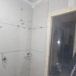 Hubert Słaboń - Wnętrze łazienki w trakcie wykańczania: ułożone płytki na ścianach z widocznymi przewodami elektrycznymi i elementami instalacji wodnej, drewniana ościeżnica drzwi.