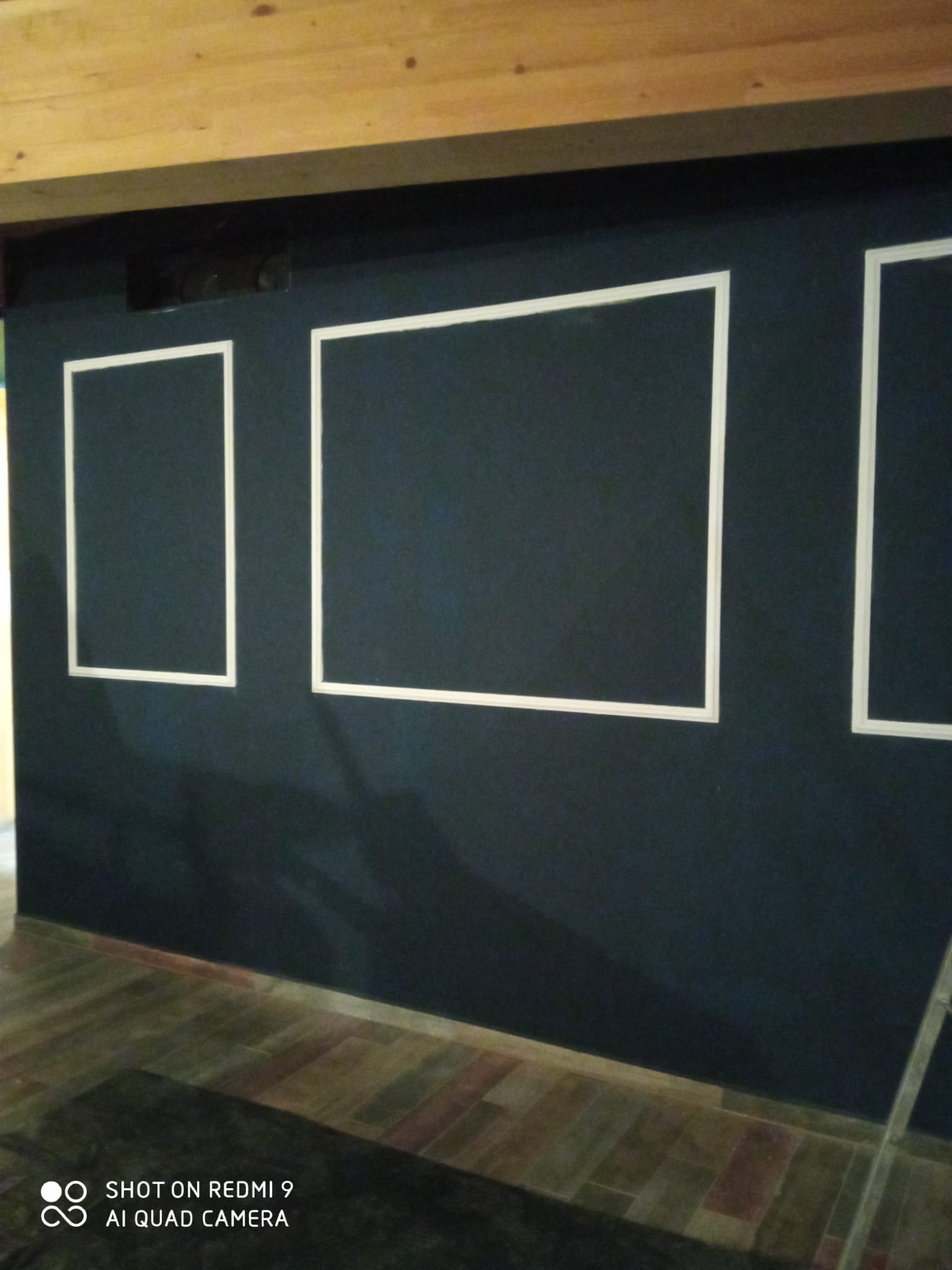 Ciemna ściana z białymi, geometrycznymi obramowaniami w kształcie prostokątów, drewniany sufit i podłoga. Widoczny fragment drabiny.