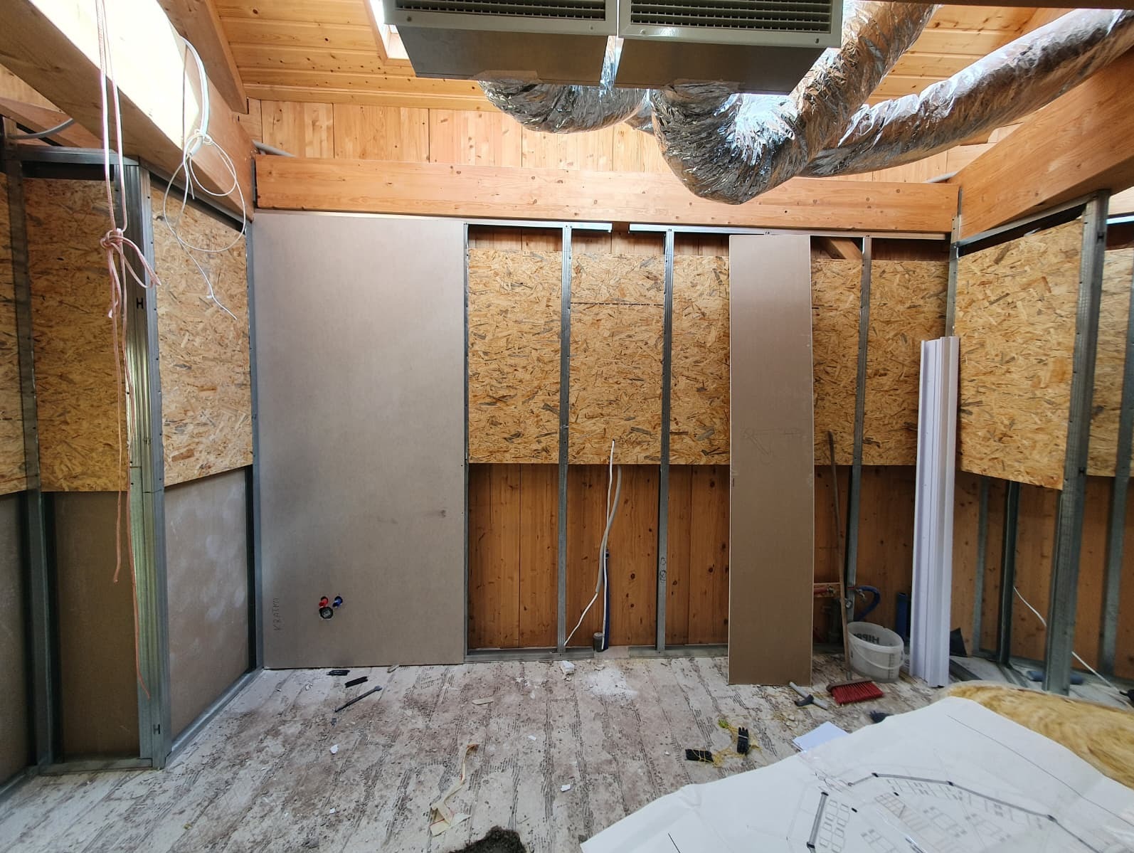 Wnętrze w trakcie remontu: metalowa konstrukcja, płyty OSB, częściowo zamontowane płyty karton-gips, widoczne instalacje i plany na podłodze. Drewniany strop z wentylacją.