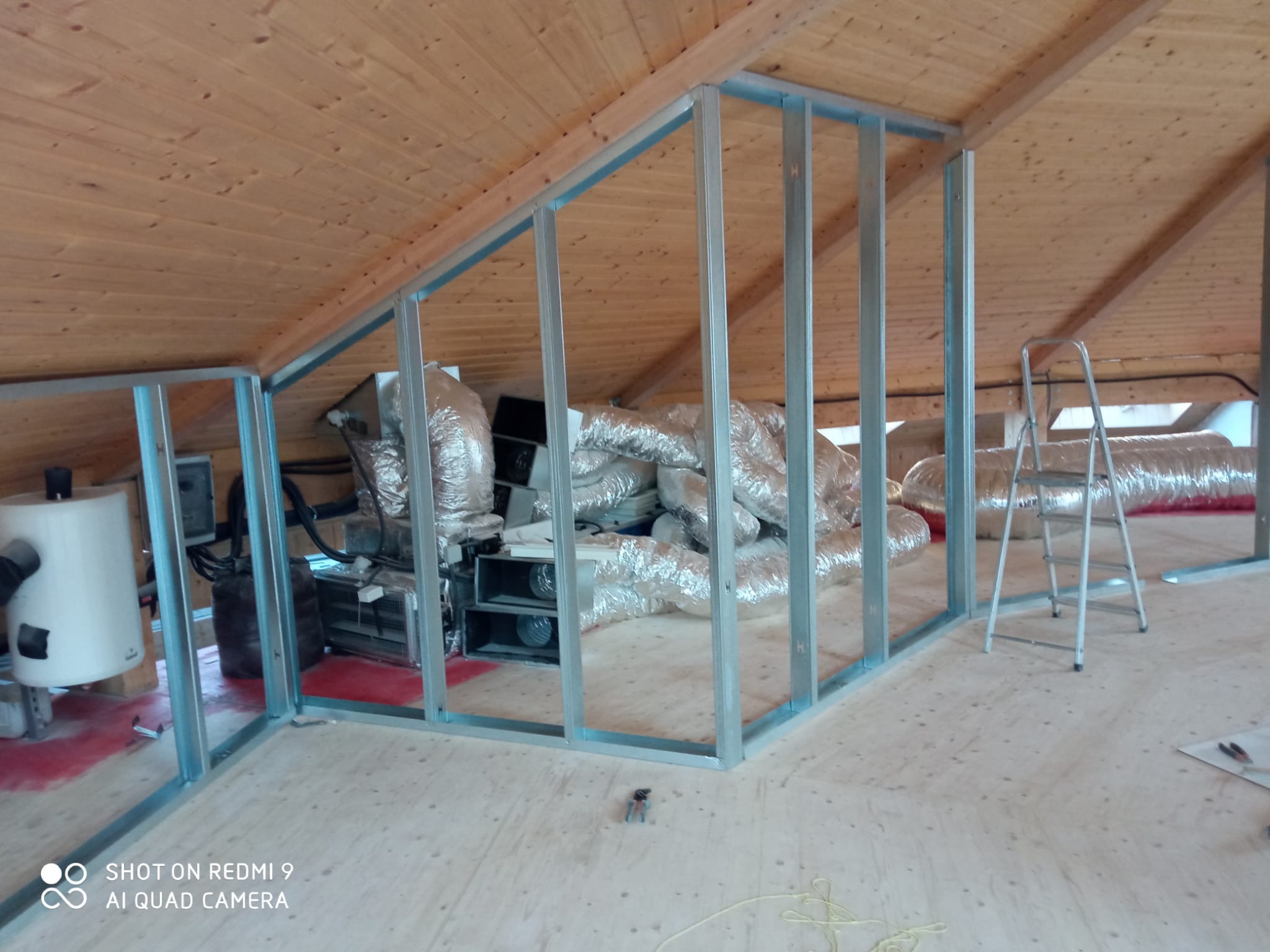 Stelaż z profili metalowych na poddaszu z drewnianą boazerią, widoczne instalacje wentylacyjne i drabina. Etap prac wykończeniowych.