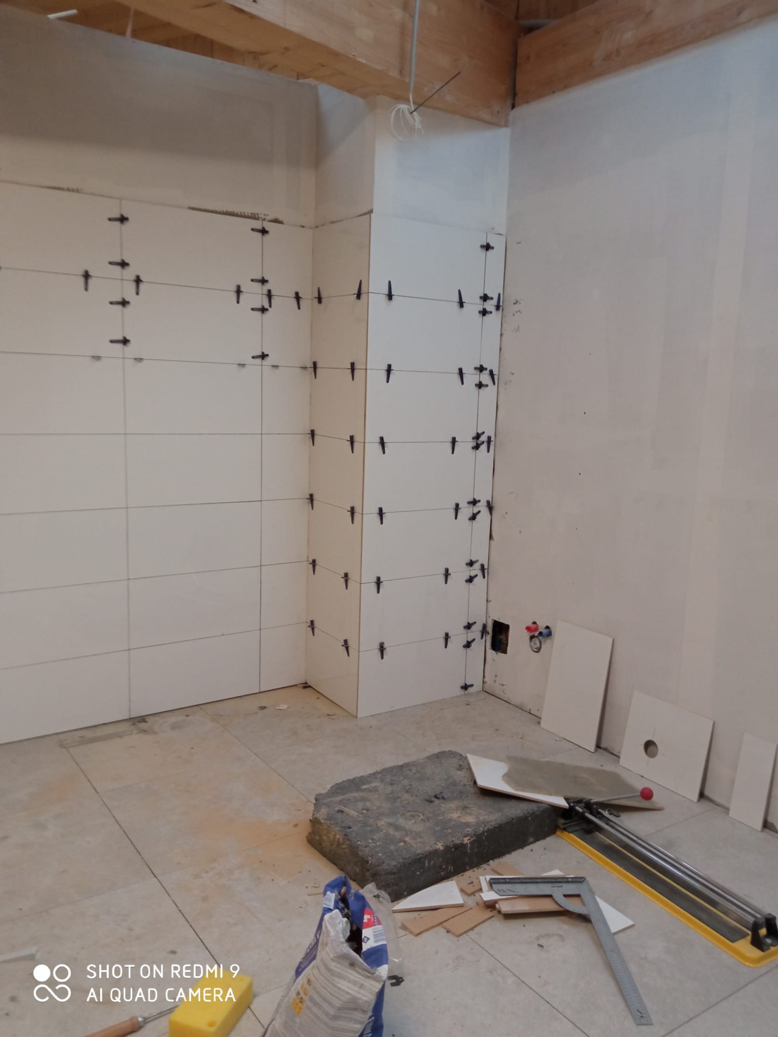 Białe płytki ścienne z krzyżykami dystansowymi w trakcie montażu w rogu pomieszczenia. Na podłodze narzędzia glazurnicze i zaprawa.