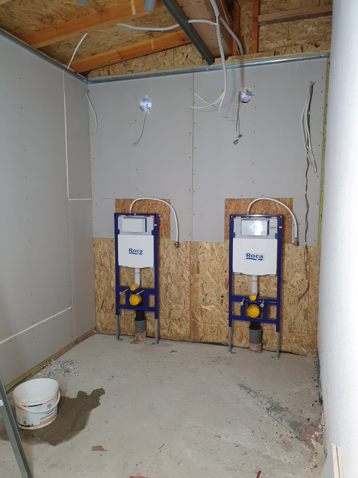 Dwa podtynkowe stelaże WC marki Roca, zamontowane na płycie OSB w surowej łazience z częściowo położonymi płytami gipsowo-kartonowymi i widoczną instalacją elektryczną.