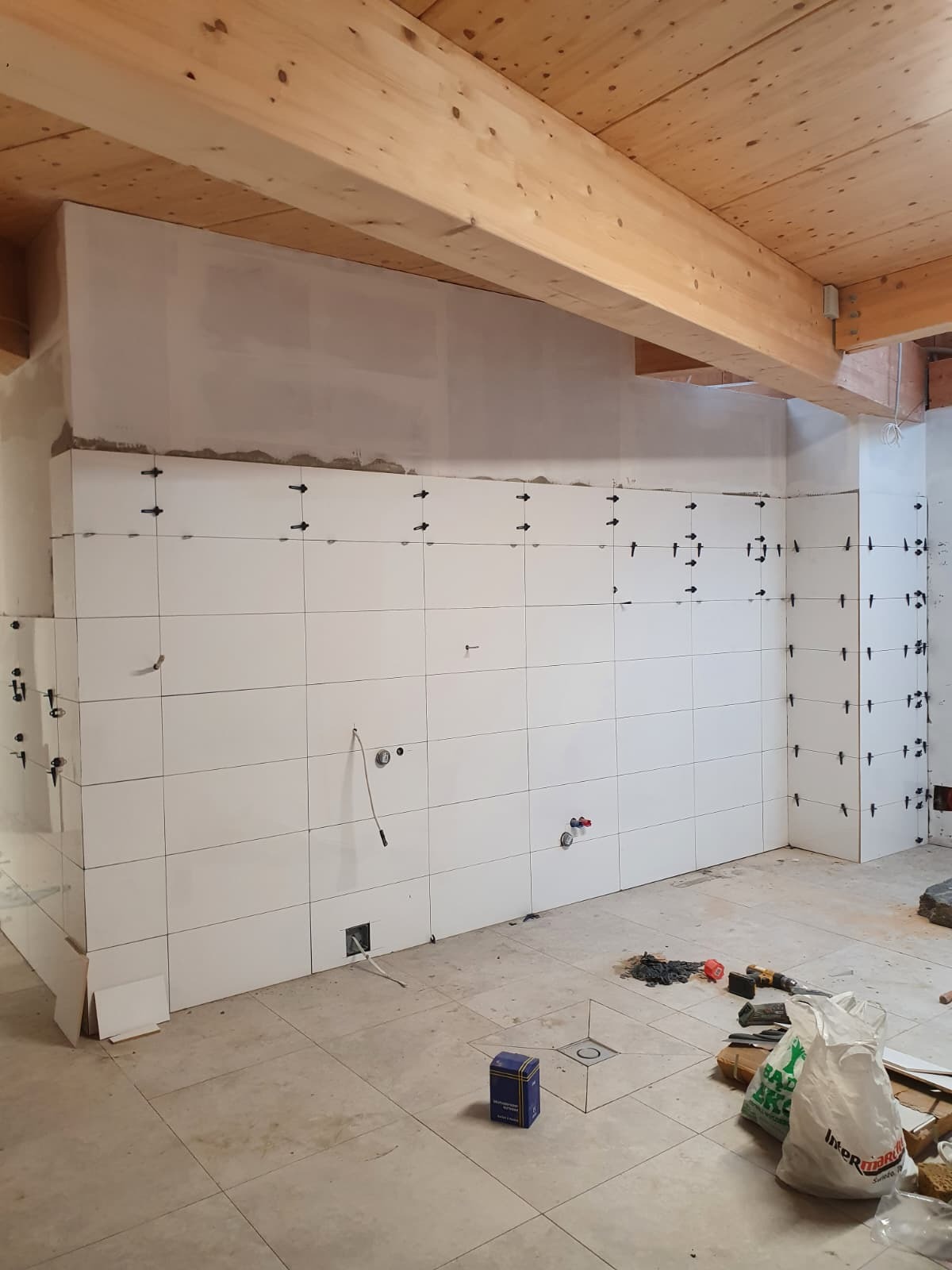 Białe płytki ścienne z krzyżykami dystansowymi w trakcie montażu, widoczne instalacje i drewniany sufit. Prace wykończeniowe w pomieszczeniu z podłogą w jasnych płytkach.