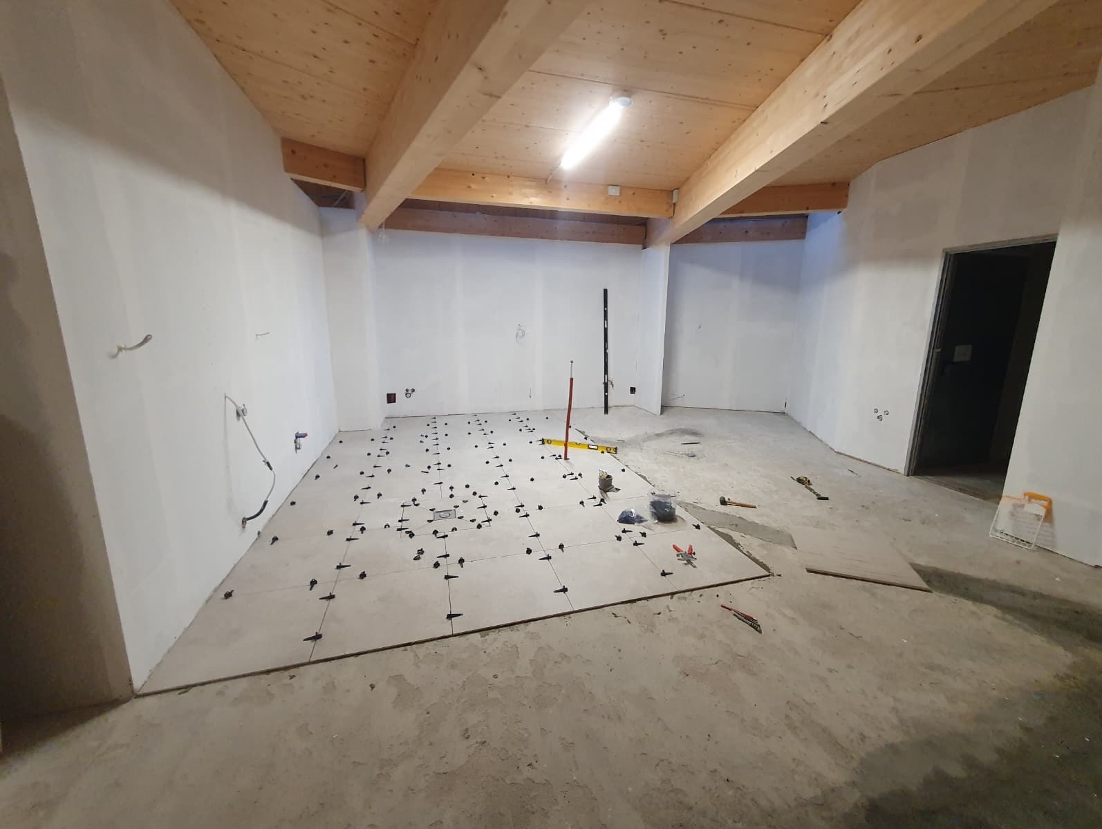 Układanie płytek podłogowych w surowym wnętrzu z drewnianym stropem. Widoczne narzędzia i krzyżyki dystansowe. Częściowo ułożona podłoga w jasnych płytkach.