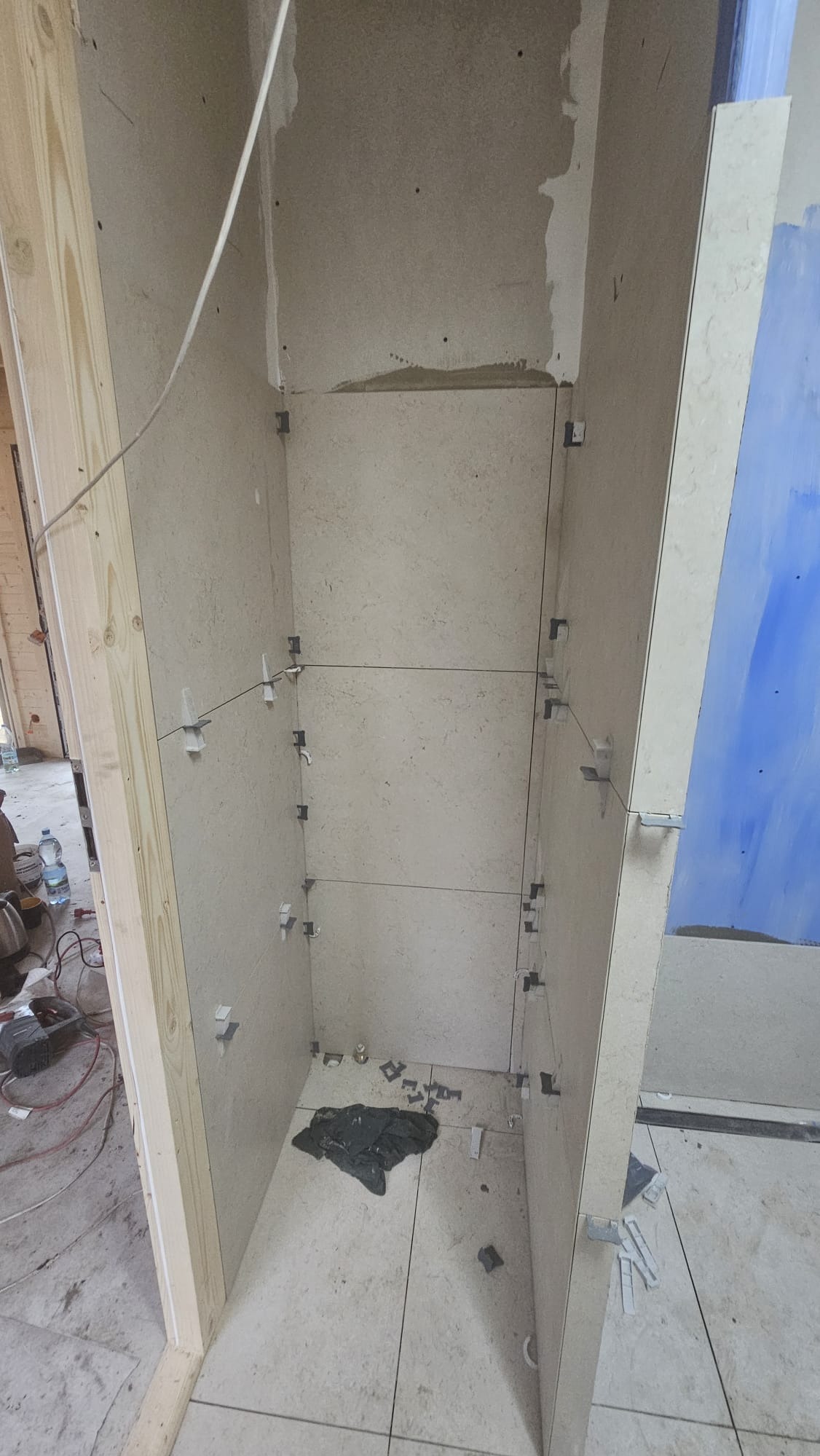 Wnęka w trakcie wykańczania płytkami, widoczne krzyżyki dystansowe i sznurki poziomujące. Częściowo ułożona podłoga z płytek. Surowy stan wykończenia ścian.