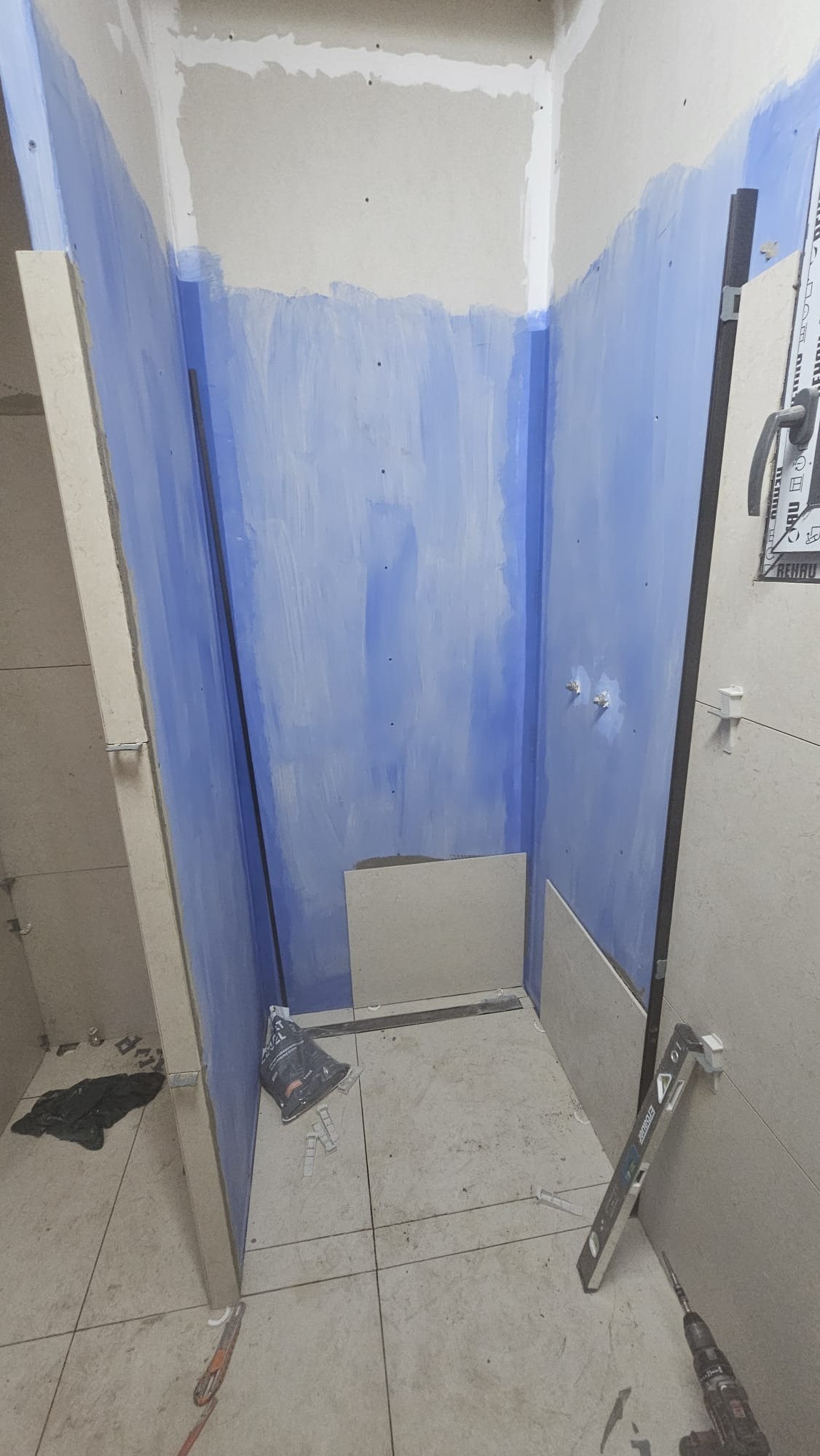 Wnętrze łazienki w trakcie wykańczania: częściowo położone płytki, ściany z niebieskim podkładem, poziomica, wiertarka i akcesoria glazurnicze.