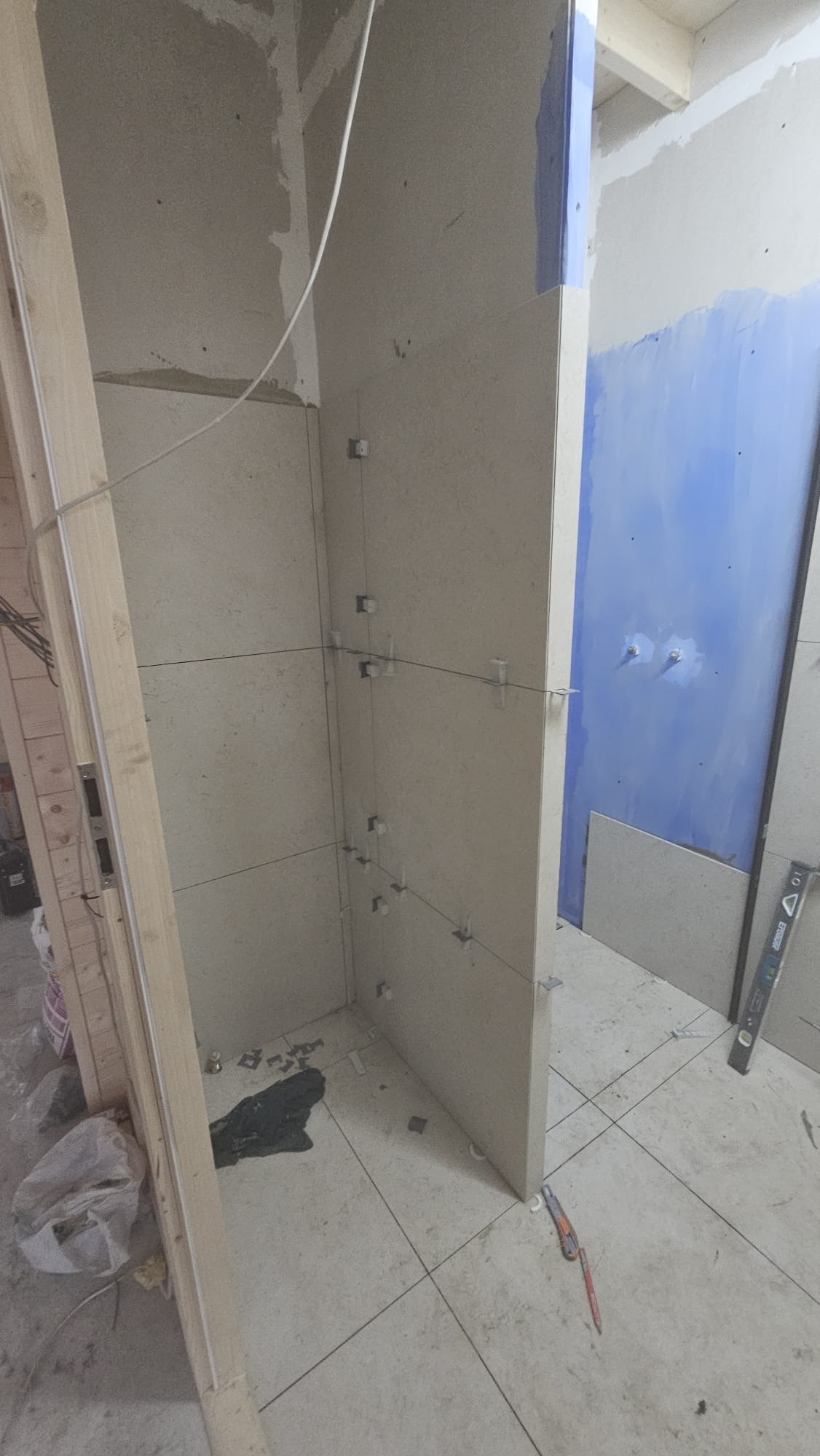 Układanie beżowych płytek w rogu łazienki, widoczne krzyżyki dystansowe i poziomica. Ściana pomalowana na niebiesko w tle. Etap prac wykończeniowych.