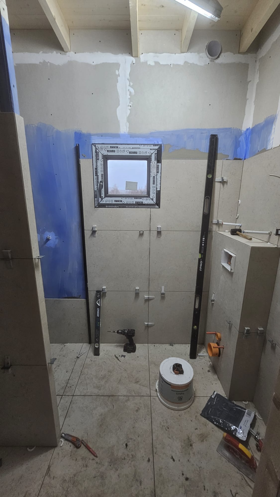 Łazienka w trakcie wykańczania: ułożone płytki na ścianach z krzyżykami dystansowymi, okno z ramą Rehau, poziomice, wiertarka i wiadro z farbą na podłodze.