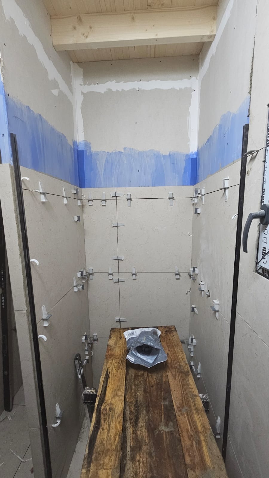 Łazienka w trakcie wykańczania: beżowe płytki ułożone na ścianach z użyciem krzyżyków dystansowych, widoczny fragment drewnianego rusztowania i niebieski pas malarski na górze.