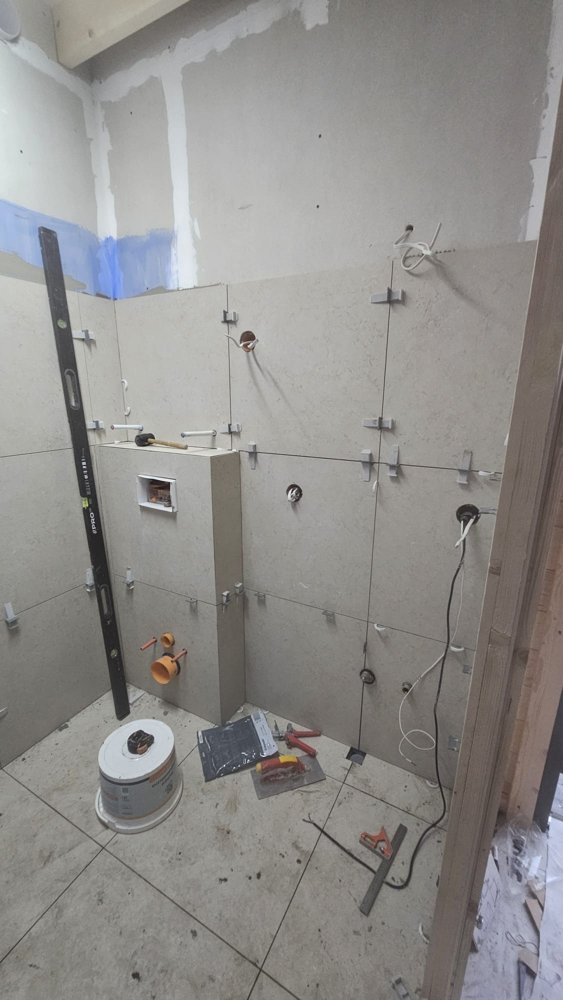 Wnętrze łazienki w trakcie układania płytek. Widoczne narzędzia, poziomica, krzyżyki dystansowe i puszki elektryczne. Ściany częściowo wykończone, sufit przygotowany do malowania.