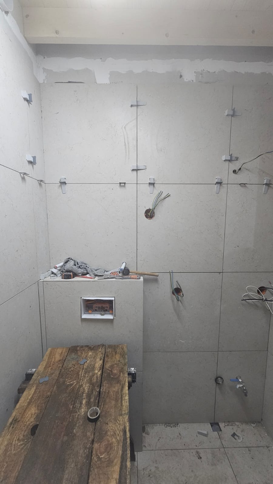 Układanie płytek w łazience, widoczne krzyżyki dystansowe i przygotowane otwory na instalacje. Na pierwszym planie drewniany stół roboczy.