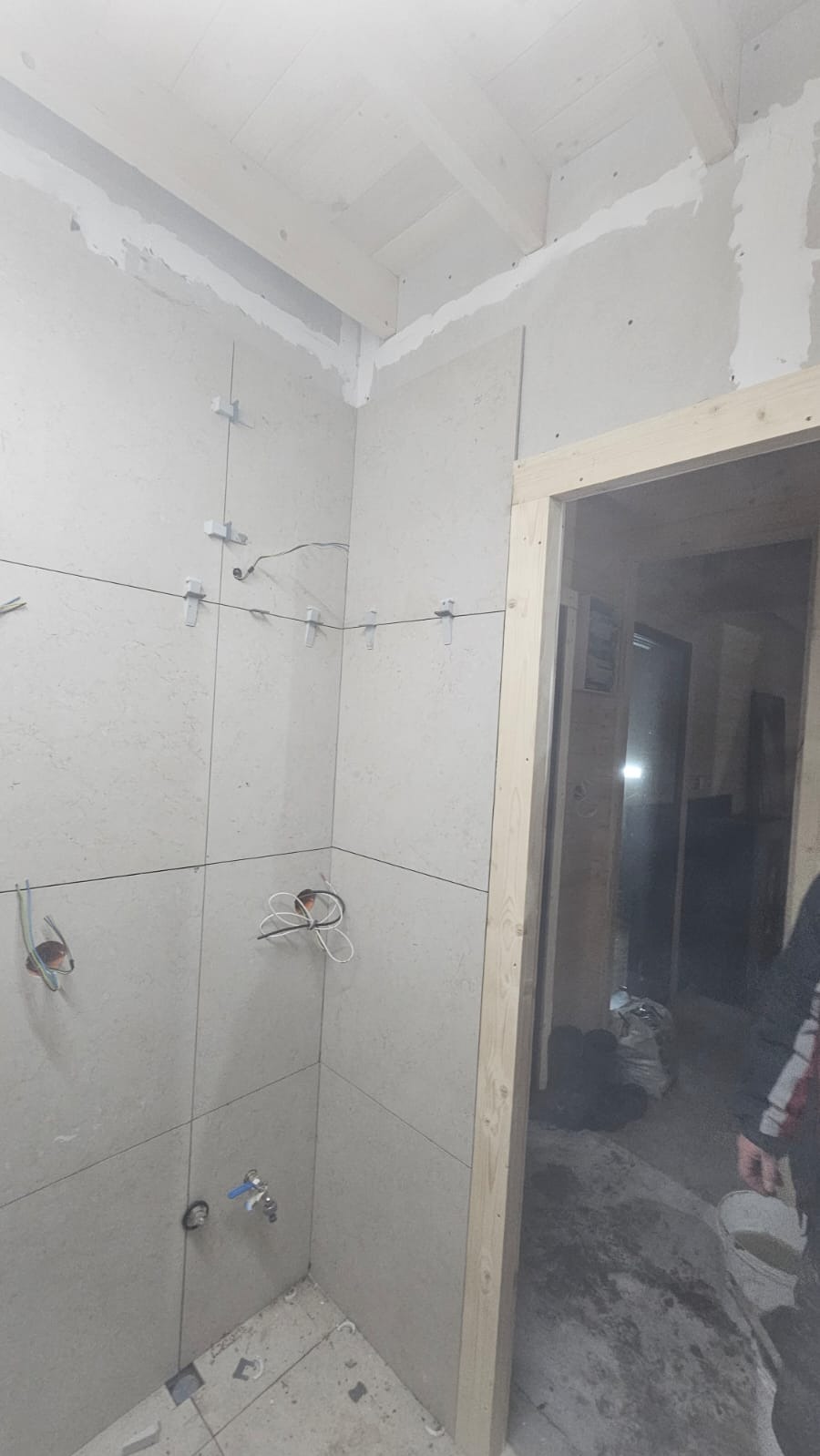 Wnętrze łazienki w trakcie wykańczania: ułożone płytki na ścianach z widocznymi przewodami elektrycznymi i elementami instalacji wodnej, drewniana ościeżnica drzwi.