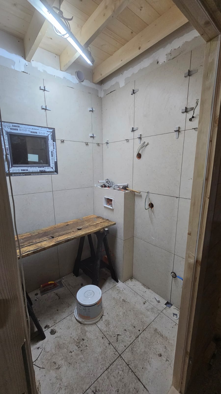 Układanie płytek w łazience w Świnoujściu. Widoczne narzędzia, stół roboczy i częściowo wykończone ściany z zamontowanymi płytkami. Sufit drewniany.