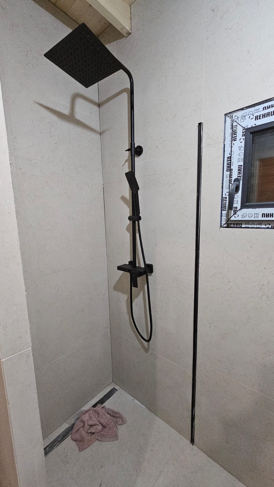 Nowoczesny, czarny zestaw prysznicowy z deszczownicą i słuchawką, zamontowany w wykończonej łazience z beżowymi płytkami i odpływem liniowym. Widoczne okno z ramą PCV.