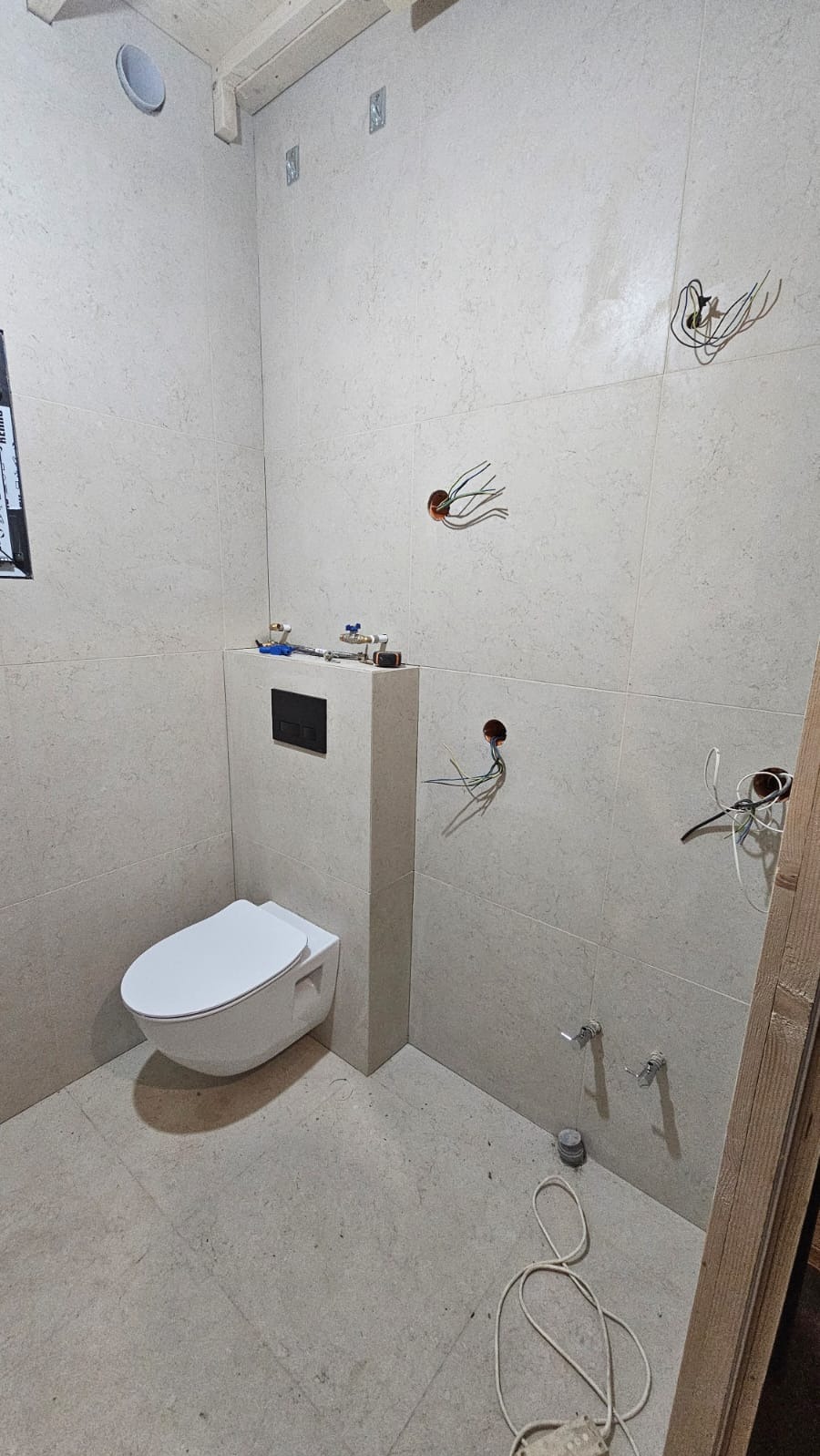 Nowoczesna, podwieszana toaleta w jasnej łazience z beżowymi płytkami. Widoczne przygotowane punkty elektryczne i hydrauliczne przed montażem armatury.