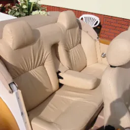 Jasna, skórzana tapicerka tylnych siedzeń nietypowego pojazdu, prawdopodobnie samochodu, z widocznymi zagłówkami i podłokietnikiem, na tle ceglanego muru i białego krzesła.