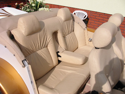 Jasna, skórzana tapicerka tylnych siedzeń nietypowego pojazdu, prawdopodobnie samochodu, z widocznymi zagłówkami i podłokietnikiem, na tle ceglanego muru i białego krzesła.