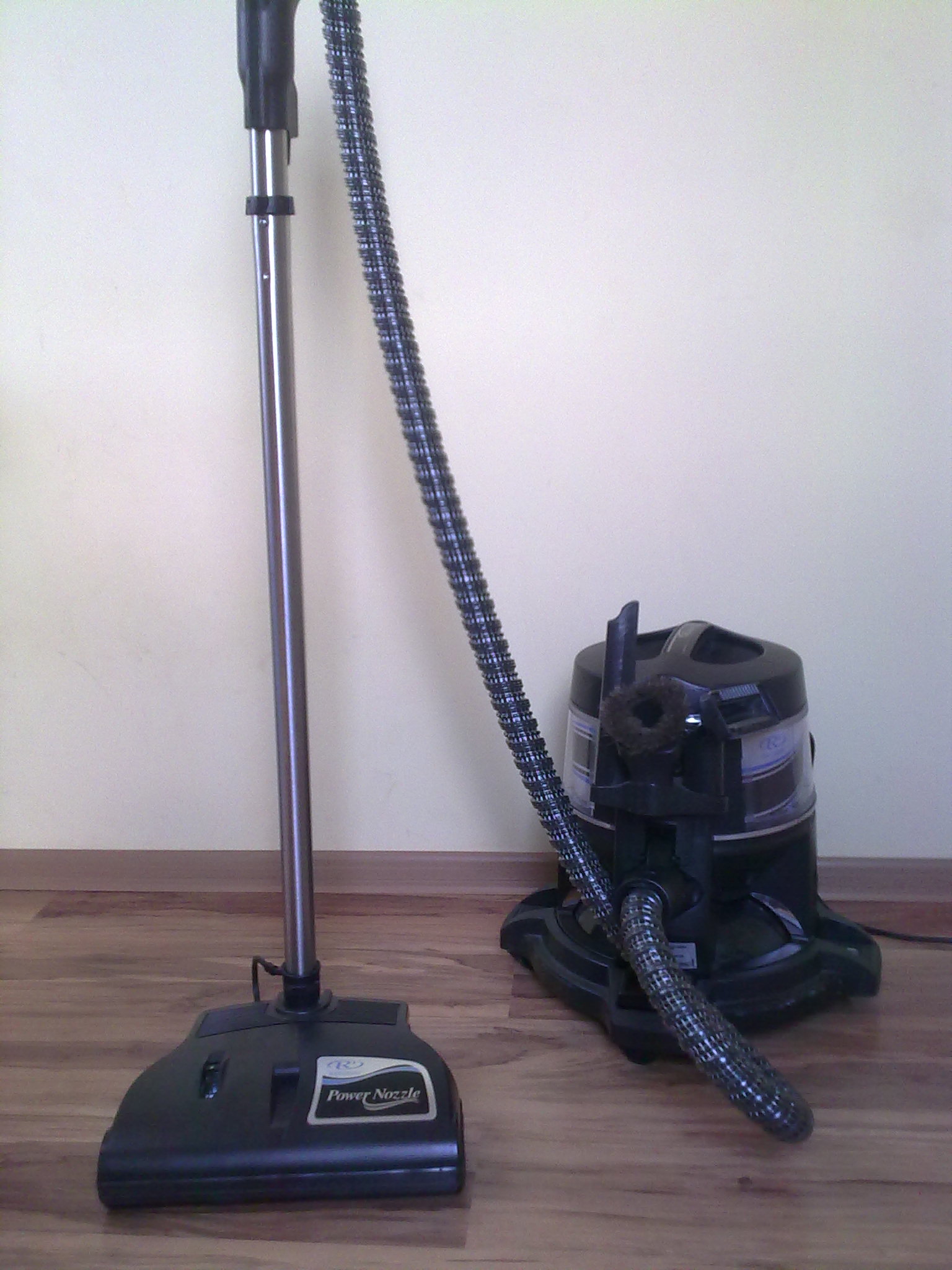 Czarny odkurzacz piorący Rainbow z nasadką Power Nozzle, gotowy do prania tapicerki w Lublewie Gdańskim, na tle drewnianej podłogi i jasnej ściany.