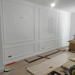 STABART - Biała ściana z dekoracyjnymi panelami w trakcie montażu. Widoczne narzędzia i zabezpieczenia podłogi. Częściowo pomalowana ściana, gniazdka elektryczne.