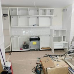 STABART - Montaż białych szafek kuchennych na tle białych płytek. Widoczne narzędzia i kartony na podłodze. Etap prac wykończeniowych w kuchni.