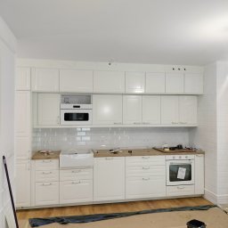 STABART - Jasna kuchnia w trakcie montażu: białe szafki z frezowanymi frontami, drewniany blat, biała cegła na ścianie. Widoczne narzędzia i zabezpieczenia podłogi.