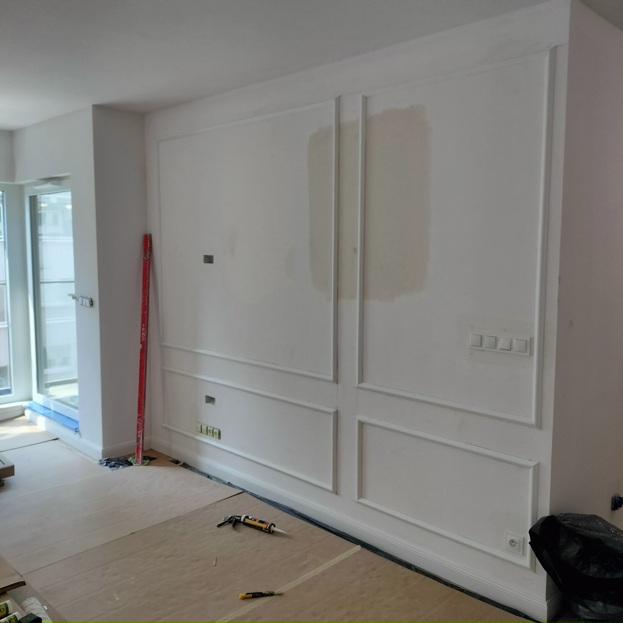 Biała ściana z dekoracyjnymi listwami w trakcie montażu. Widoczne narzędzia, poziomica i częściowo zakryta podłoga. W tle okno i fragment pomieszczenia.