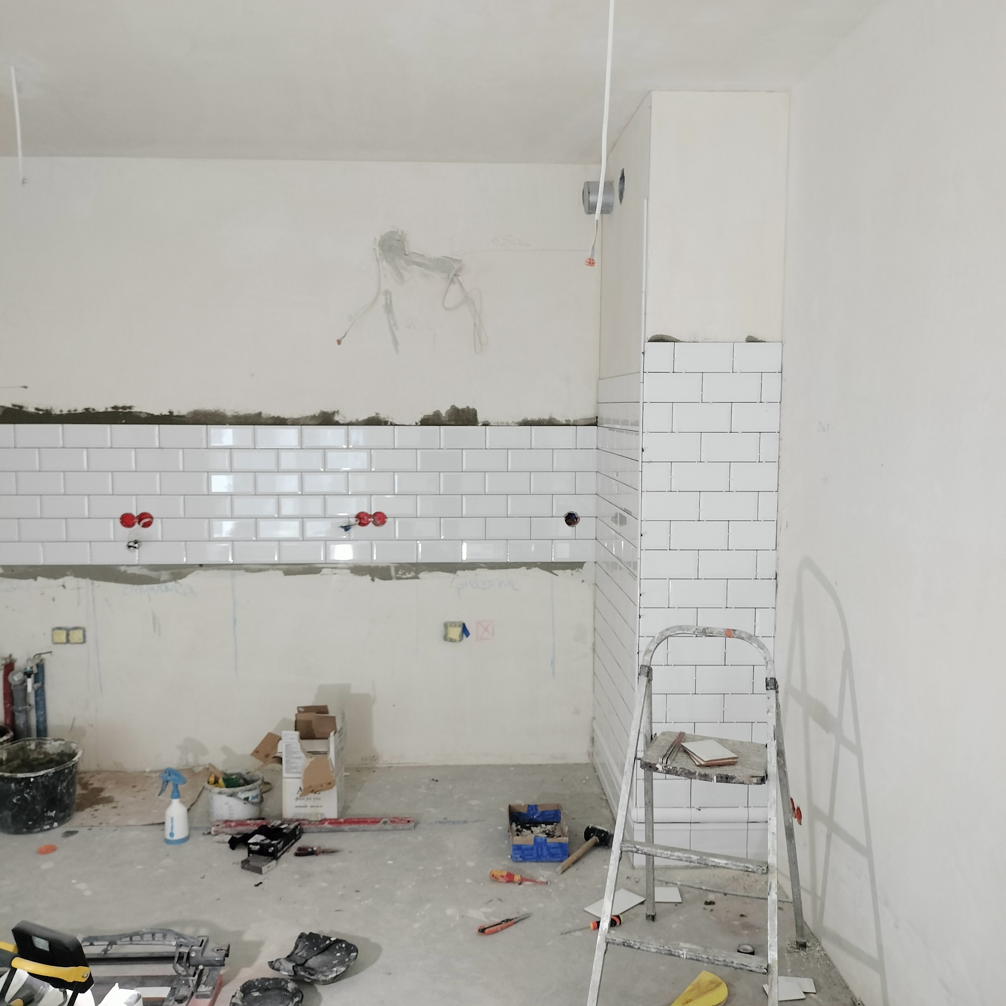 Białe, prostokątne płytki ułożone na ścianie do połowy wysokości, widoczne narzędzia glazurnicze i drabina. Prace w toku, surowy stan wykończenia.