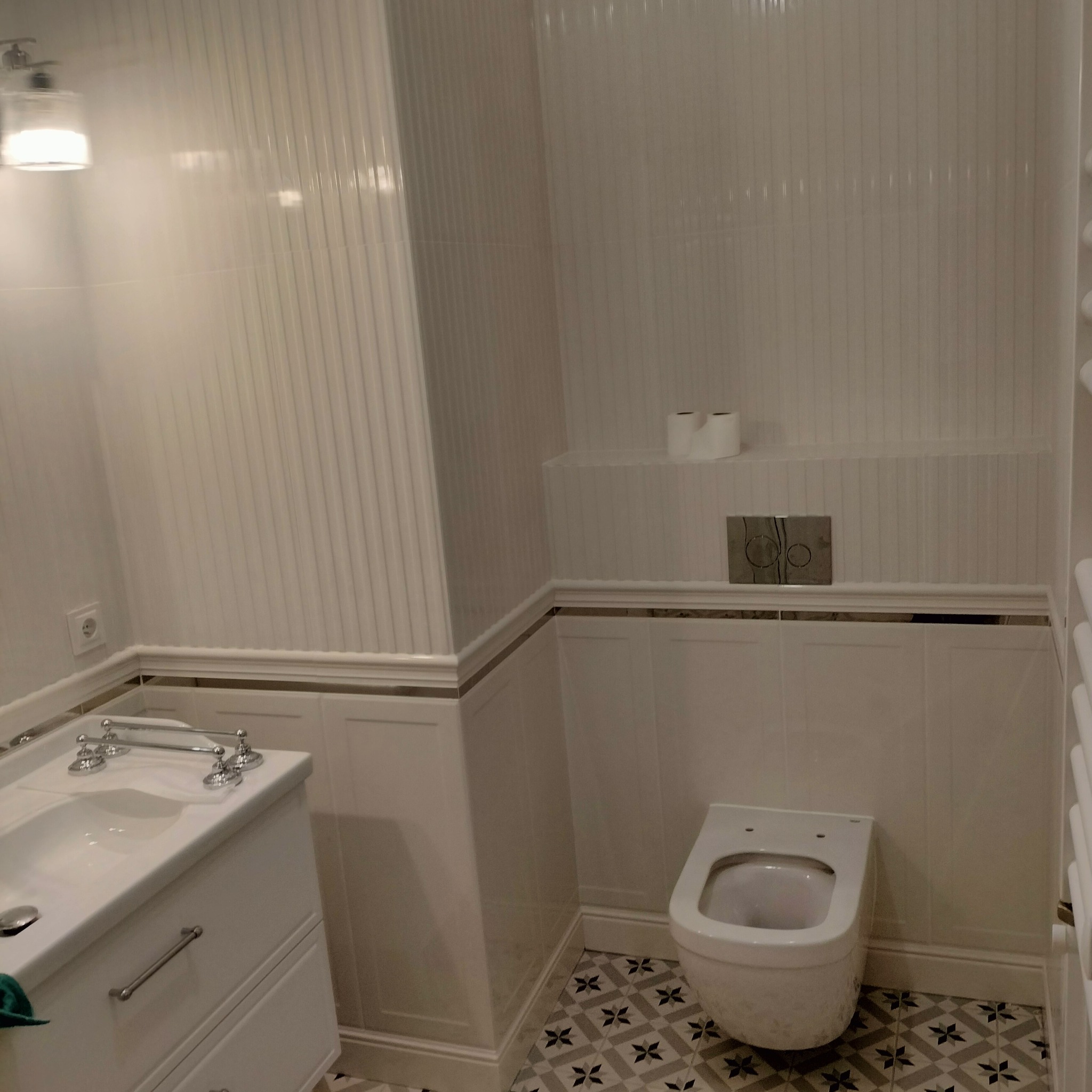 Elegancka, wykończona łazienka w bieli z dekoracyjnymi płytkami podłogowymi. Umywalka z szafką i chromowaną armaturą, wisząca toaleta. Ściany wyłożone płytkami z subtelnym wzorem.