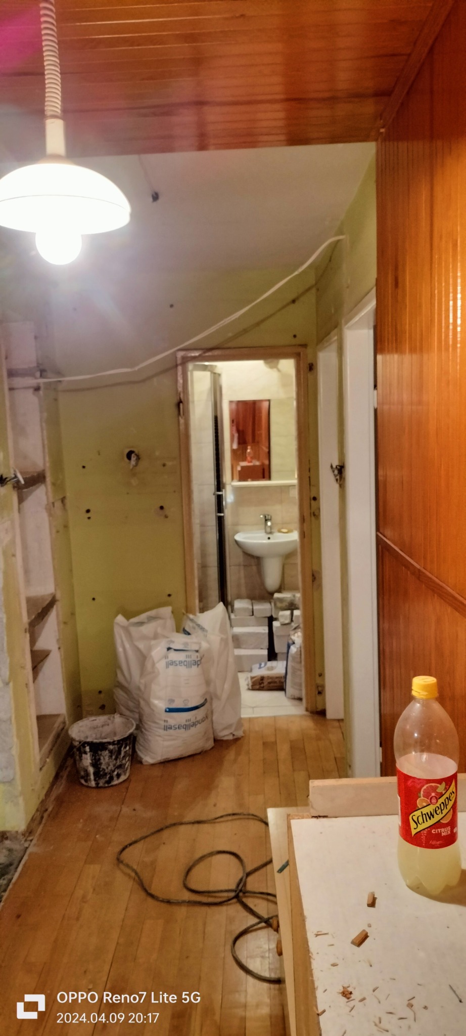 Wnętrze w trakcie remontu: widoczna łazienka z umywalką, worki z zaprawą, wiadro i narzędzia. Drewniana podłoga i boazeria. Etap przygotowawczy do prac wykończeniowych.