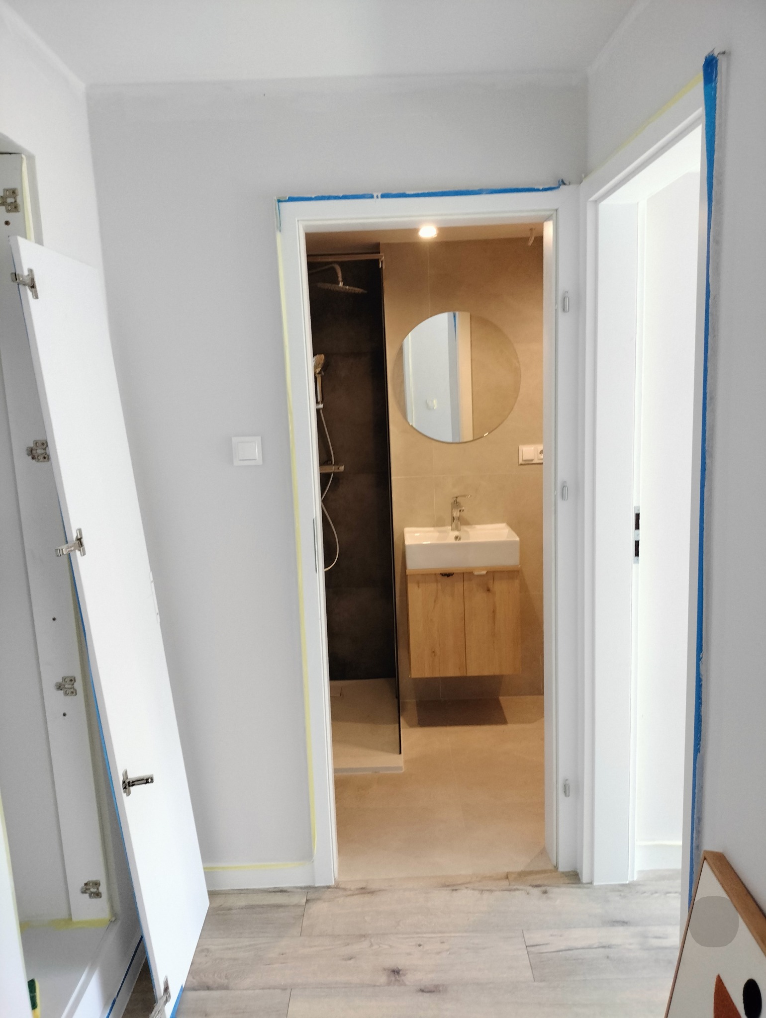 Widok na wykończoną łazienkę z kabiną prysznicową i szafką z umywalką, przez otwarte drzwi z zabezpieczeniami malarskimi. Drzwi do łazienki częściowo zdjęte.