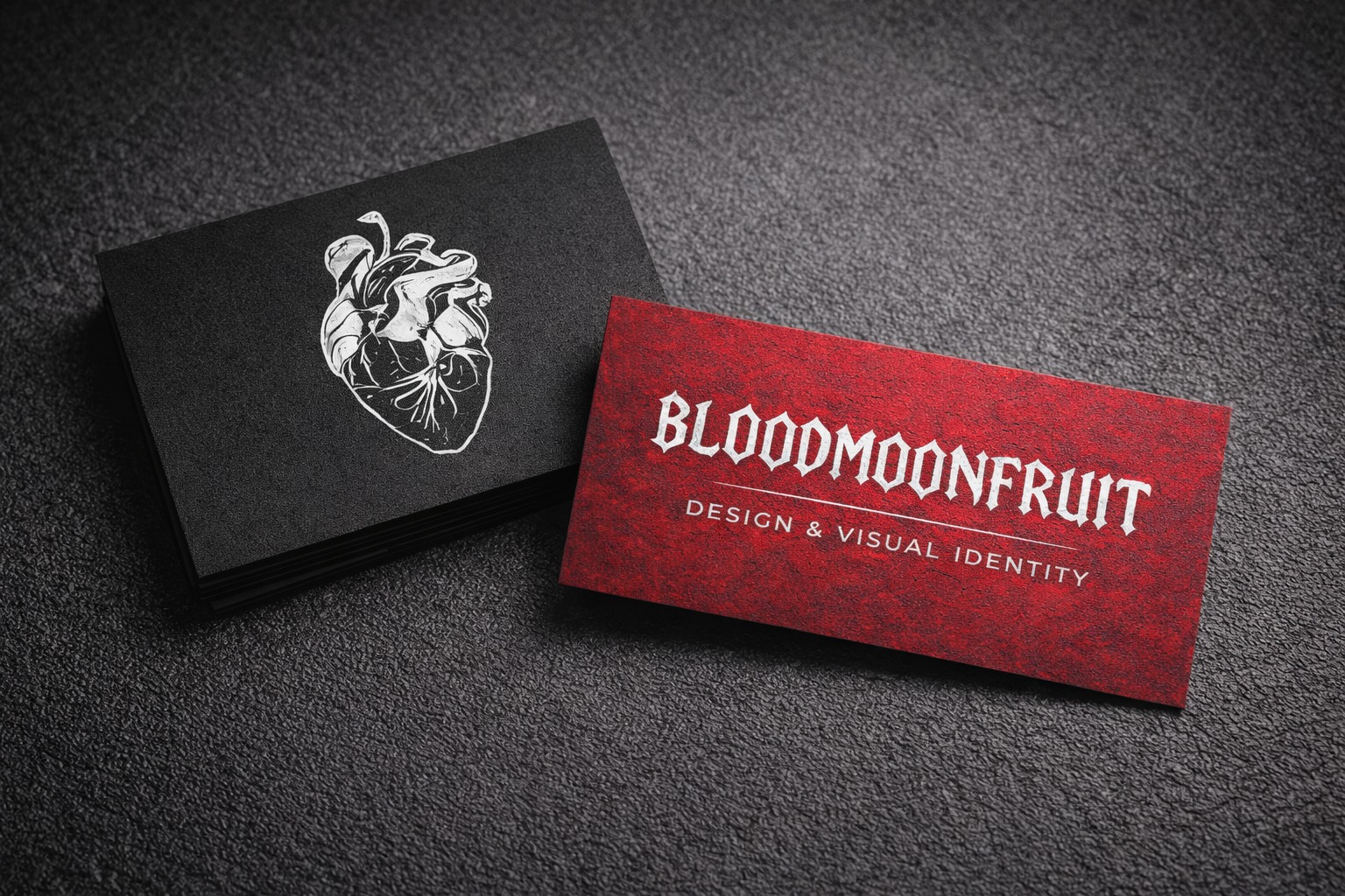 Dwie wizytówki: czarna z grafiką serca i czerwona z napisem 'BLOODMOONFRUIT DESIGN & VISUAL IDENTITY', leżące na teksturowanym, ciemnym tle.