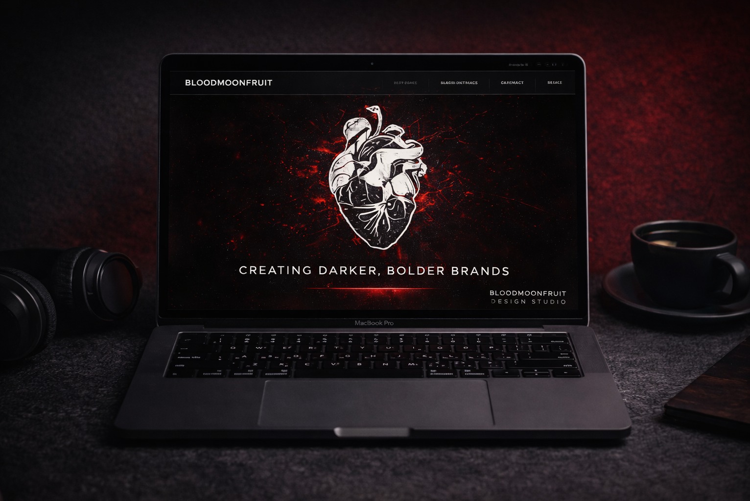 Laptop z otwartą stroną internetową 'Bloodmoonfruit Design Studio' z grafiką serca i hasłem 'Creating Darker, Bolder Brands'. Obok słuchawki i filiżanka kawy.