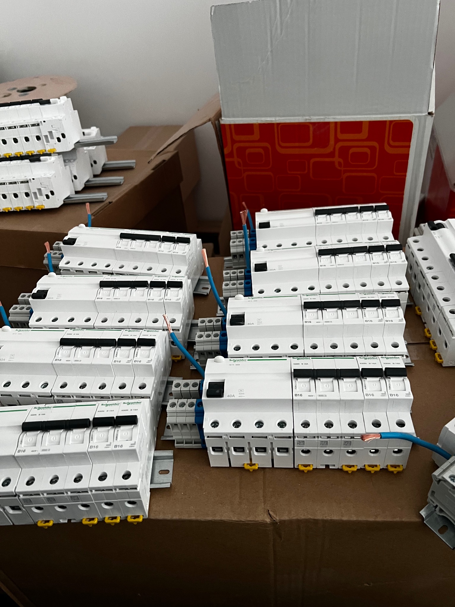 Przygotowane do montażu wyłączniki nadprądowe Schneider Electric B16 40A, ułożone w rzędach na kartonowym podłożu, z podłączonymi przewodami. Widoczne elementy instalacji.