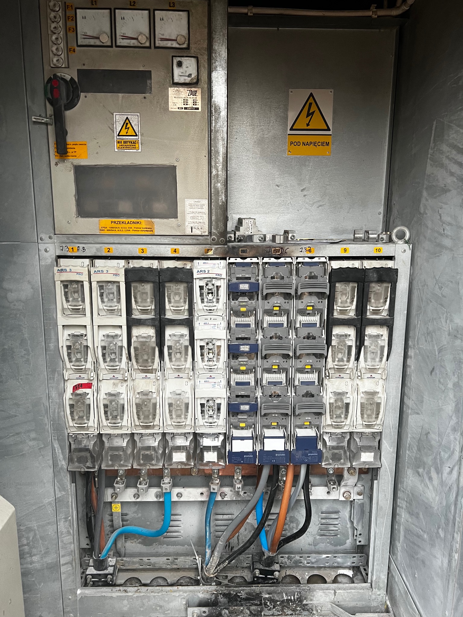 Rozdzielnica elektryczna z bezpiecznikami i przewodami, widoczne oznaczenia i tabliczki ostrzegawcze. Elementy instalacji w szarej obudowie.
