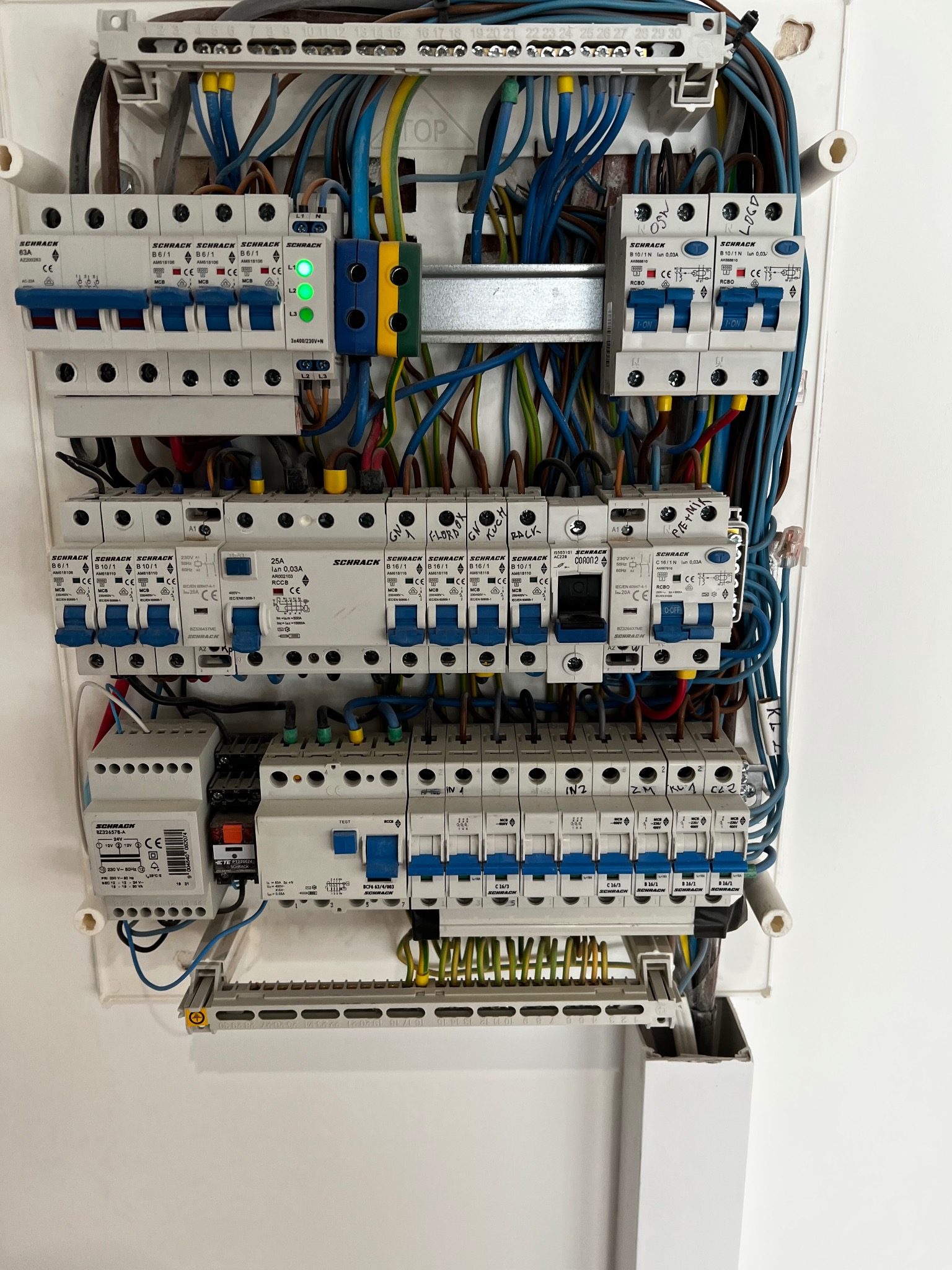 Rozdzielnica elektryczna Schrack z bezpiecznikami, przewodami w kolorach niebieskim, żółtym i brązowym. Widoczne oznaczenia na elementach i zielone diody sygnalizacyjne.