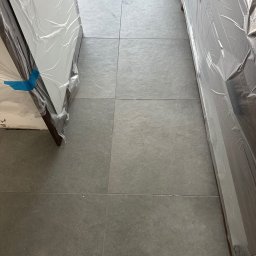 ADIKRISBAU Remonty i Wykończenia Krzysztof Smaza - Szare płytki podłogowe z fugami, częściowo zakryte folią ochronną, prawdopodobnie zabezpieczającą nowe meble lub drzwi przed zabrudzeniem podczas prac wykończeniowych.