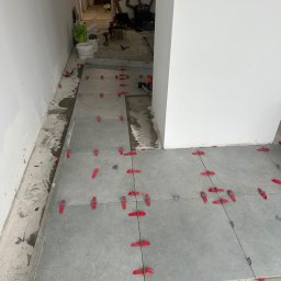 ADIKRISBAU Remonty i Wykończenia Krzysztof Smaza - Układanie szarych płytek podłogowych z czerwonymi i szarymi klipsami poziomującymi w trakcie prac wykończeniowych. Widoczne narzędzia i materiały budowlane w tle.